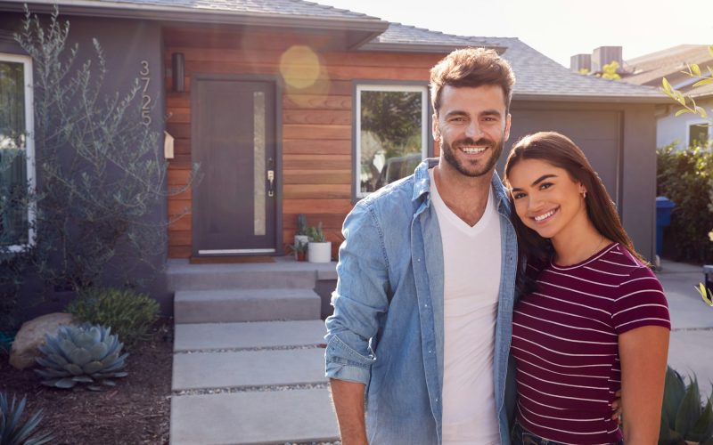 Portrait Of Couple Standing Outdoors In Front Of House With For Sale Sign In Garden